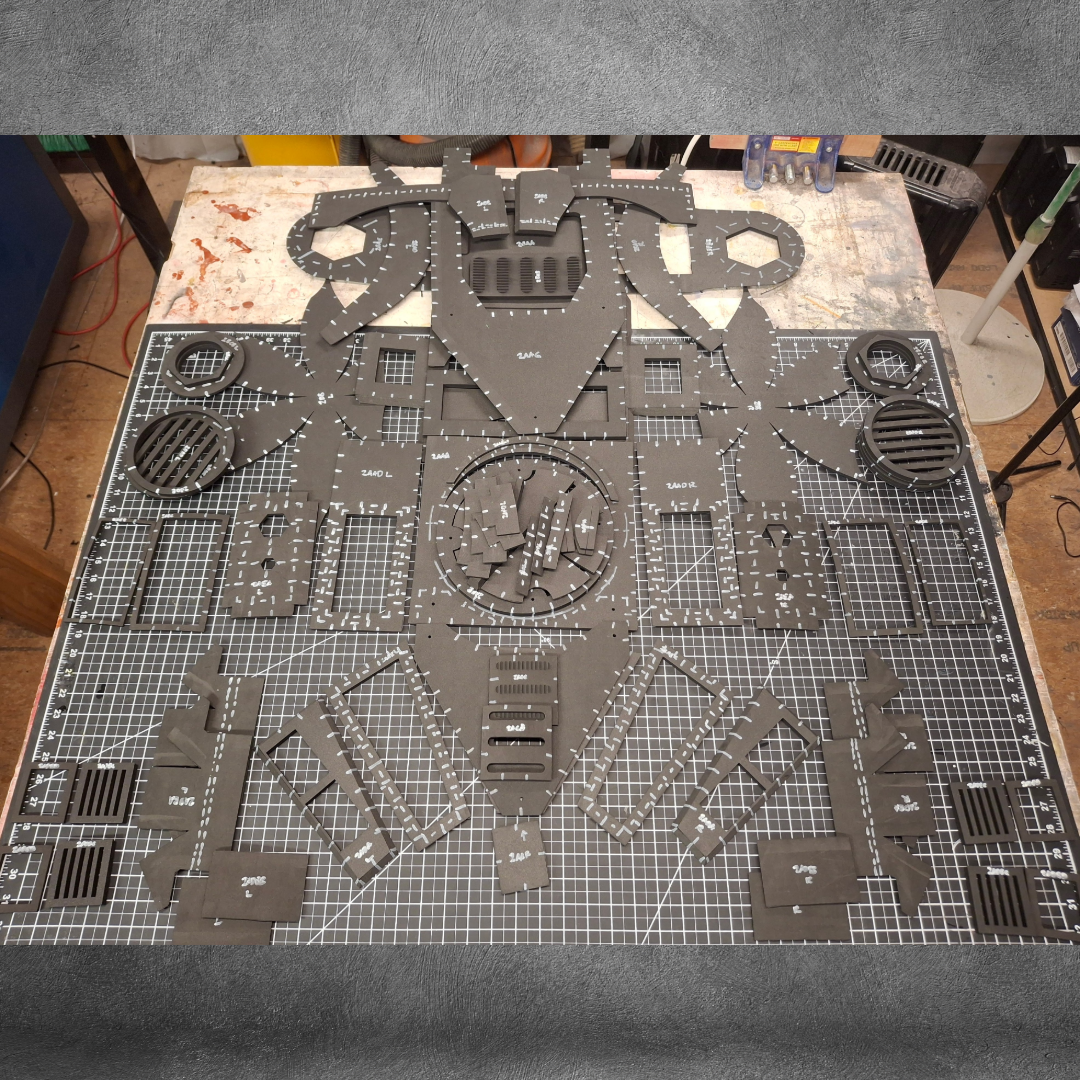 Black foam board pieces, each labeled and precisely cut using advanced EVA foam crafting techniques, are laid out on a gridded cutting mat, forming parts for a model assembly—specifically the V.10 Tactical Backpack Pattern project. Tools and workshop items are visible around the mat on a wooden workspace.