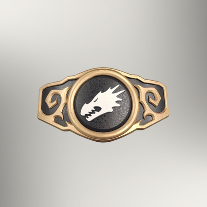 A gold-toned metal belt buckle featuring a central circular black insert with a white dragon head emblem, flanked by ornate scrollwork on a light gray gradient background.
