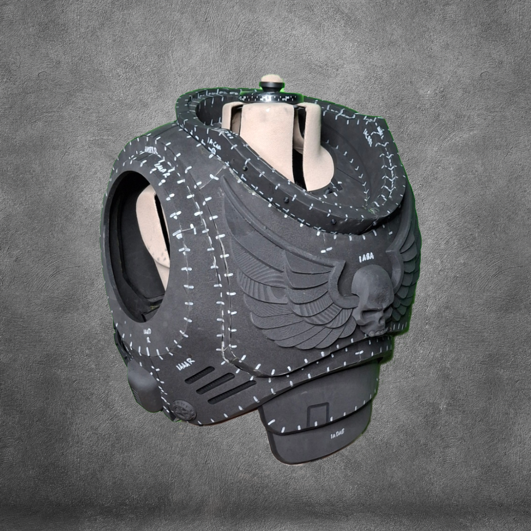 A black, futuristic helmet-like object featuring winged skull designs and white stitched patterns sits against a gray textured background, showcasing the unique style of the V.10 Tactical Chest Pattern.
