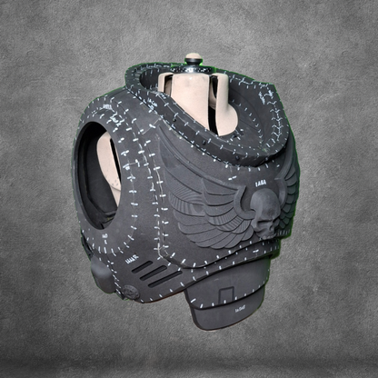 A black, futuristic helmet-like object featuring winged skull designs and white stitched patterns sits against a gray textured background, showcasing the unique style of the V.10 Tactical Chest Pattern.