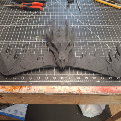 A 3D-printed black dragon head with outstretched wings sits on a gridded work surface, highlighting the intricate details from the V.10 Fire Lizards Chest Detail (3D Print | Files Only). Various tools and a ruler are visible in the background.