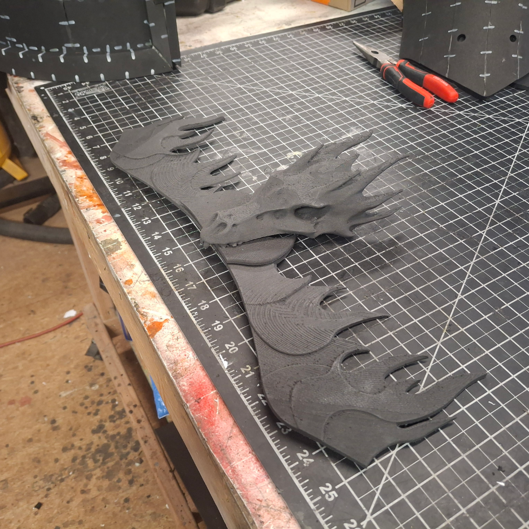 A 3D-printed black dragon head and wings—featuring intricate details from the V.10 Fire Lizards Chest Detail (3D Print | Files Only)—rest on a cutting mat, with tools and crafting materials visible in the background.