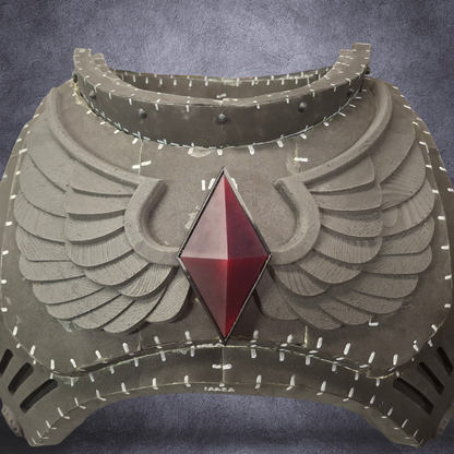 A fantasy-style winged armor chest plate or helmet front piece in weathered gray metal, featuring a large faceted red gemstone at the center and white stitched detailing across the surface, shown against a dark textured gray background.