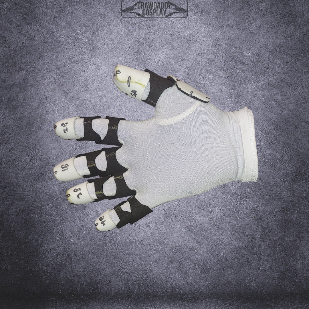 A white glove with black straps is shown palm-down on a textured gray surface, with handwritten numbers marking each finger segment—making it the ideal reference for using the Galactic Warriors Universal Glove Pattern digital download as an EVA foam template for your next project.
