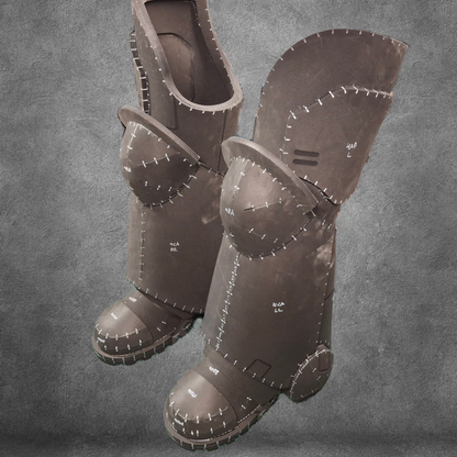 Two large, brown, foam costume boots featuring exaggerated stitched seams and armor-inspired panels—perfect for finishing your Galactic Warriors legs—stand upright against a textured gray background. The V.10 Tactical Legs Pattern EVA foam template is available as a convenient digital download.