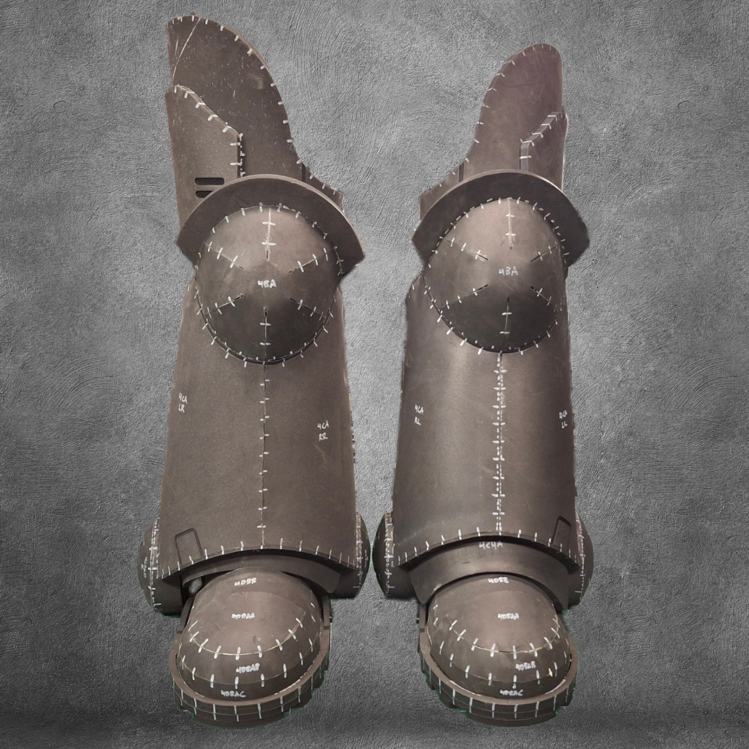 A pair of V.10 Tactical Legs Pattern black metal greaves featuring white markings and measurement lines, inspired by Galactic Warriors legs, displayed against a gray textured background.