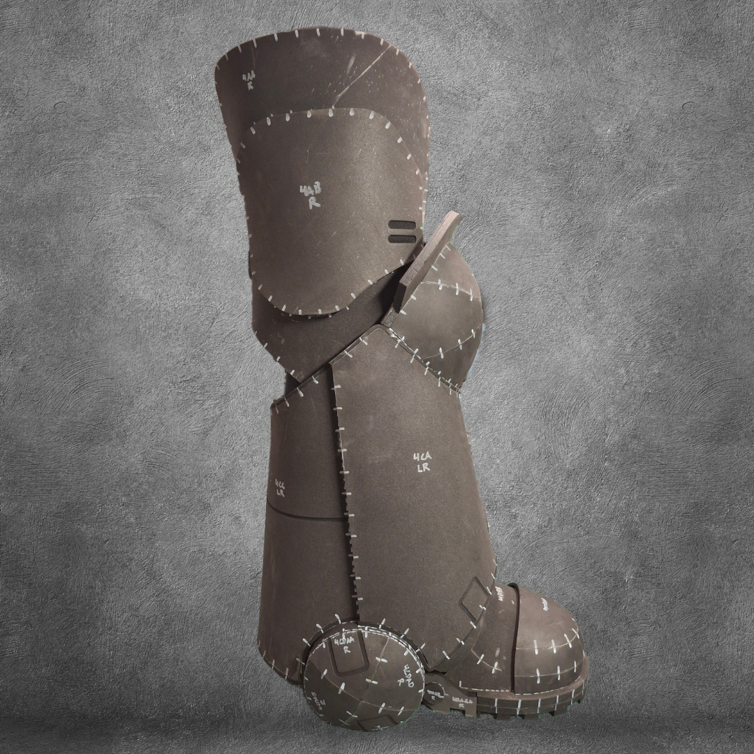 A large brown boot, stitched together with visible seams and markings, stands upright against a gray textured backdrop—resembling a patchwork sculpture—ideal as the V.10 Tactical Legs Pattern EVA foam template for crafting Galactic Warriors legs, available as a digital download.
