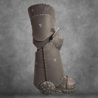 A large brown boot, stitched together with visible seams and markings, stands upright against a gray textured backdrop—resembling a patchwork sculpture—ideal as the V.10 Tactical Legs Pattern EVA foam template for crafting Galactic Warriors legs, available as a digital download.