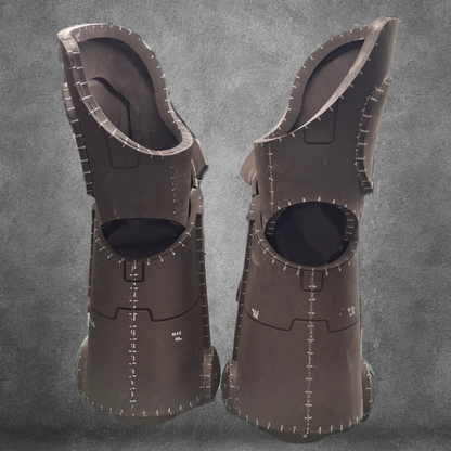 Two tall, black, segmented foam costume boots with visible seams and markings—perfect as Galactic Warriors legs—stand upright on a gray textured background. Ideal for cosplay, these boots can be recreated using the V.10 Tactical Legs Pattern EVA foam template, available as a digital download.