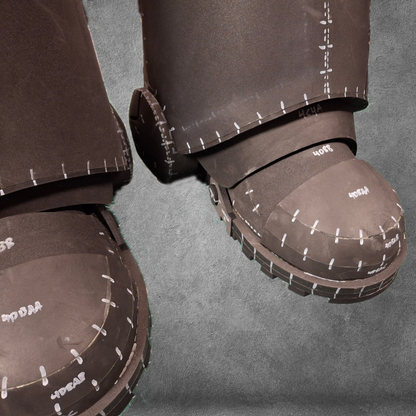 Two large costume boots, modeled after the V.10 Tactical Legs Pattern and crafted from dark EVA foam with visible seams and labeled pattern markings, are displayed against a gray background—perfect for use as an EVA foam template inspired by Galactic Warriors legs.