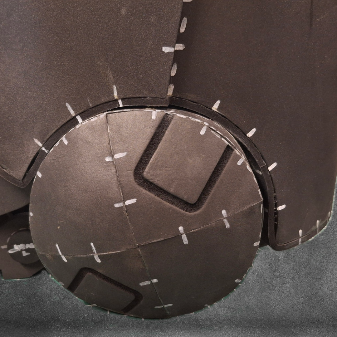 A close-up of the V.10 Tactical Legs Pattern reveals a round, segmented design with dark panels and white dashed lines, reminiscent of sci-fi armor or a robot’s joint—specifically inspired by Galactic Warriors legs. Set against a gray, textured background, this digital download is perfect for crafting an EVA foam template.