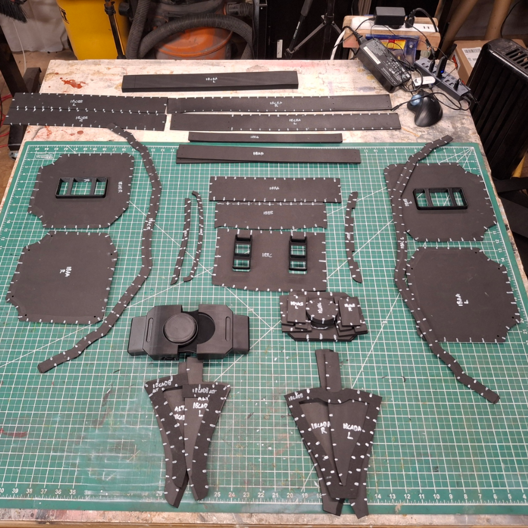 Foam costume armor pieces with labeled edges are arranged on a green cutting mat in a workshop, ready for assembly. Showcasing the V.10 Tactical Waist Pattern, this digital download provides precise guidance for crafting your own detailed costume waist armor. Tools and supplies can be seen in the background on shelves and the workbench.