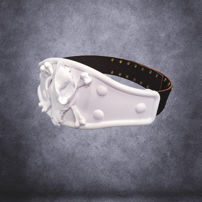 A white padded collar or headband with a sculpted skull-and-crossbones motif on the front, attached to a dark brown strap with brass-tone studs, shown against a textured gray studio background.